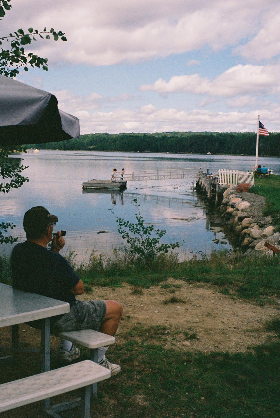 A man sits at a picnic table looking out at a lake with a dock and two children.