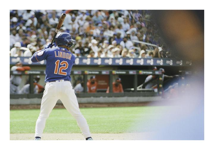 A baseball player in a blue jersey with the name "LINDOR" and number "12" on the back is at bat, facing away from the viewer, with spectators in the stands behind him.