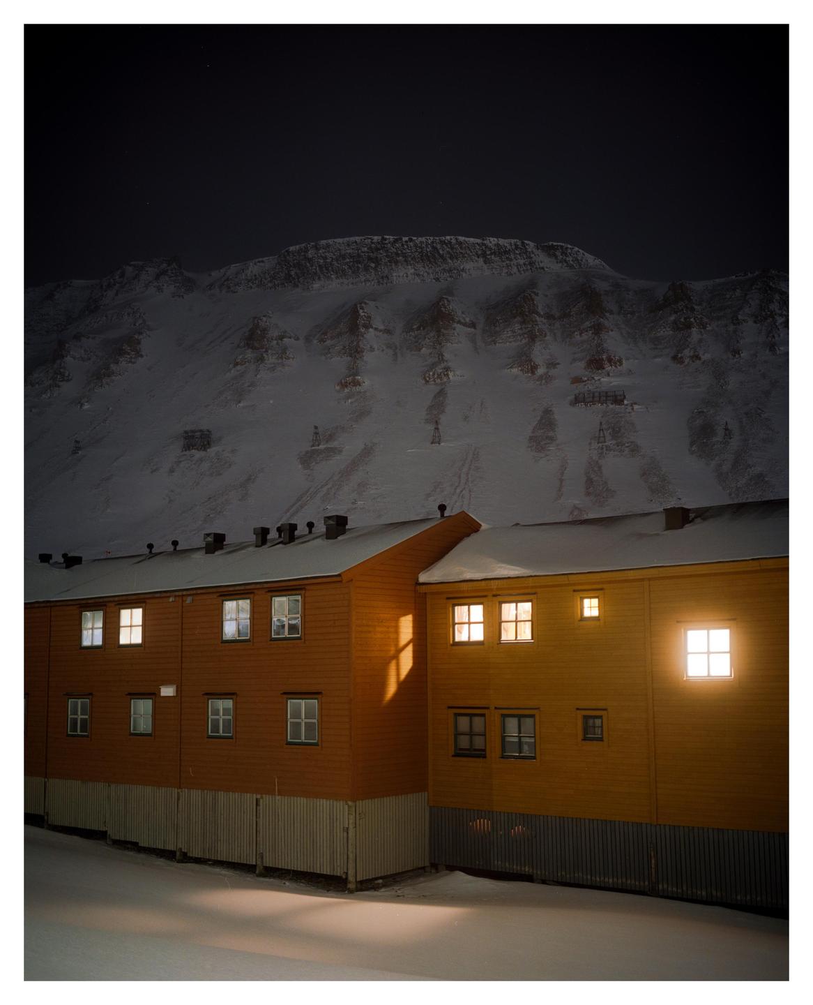 A yellow building with illuminated windows stands in the snow at night, with a large snow-covered mountain in the background.