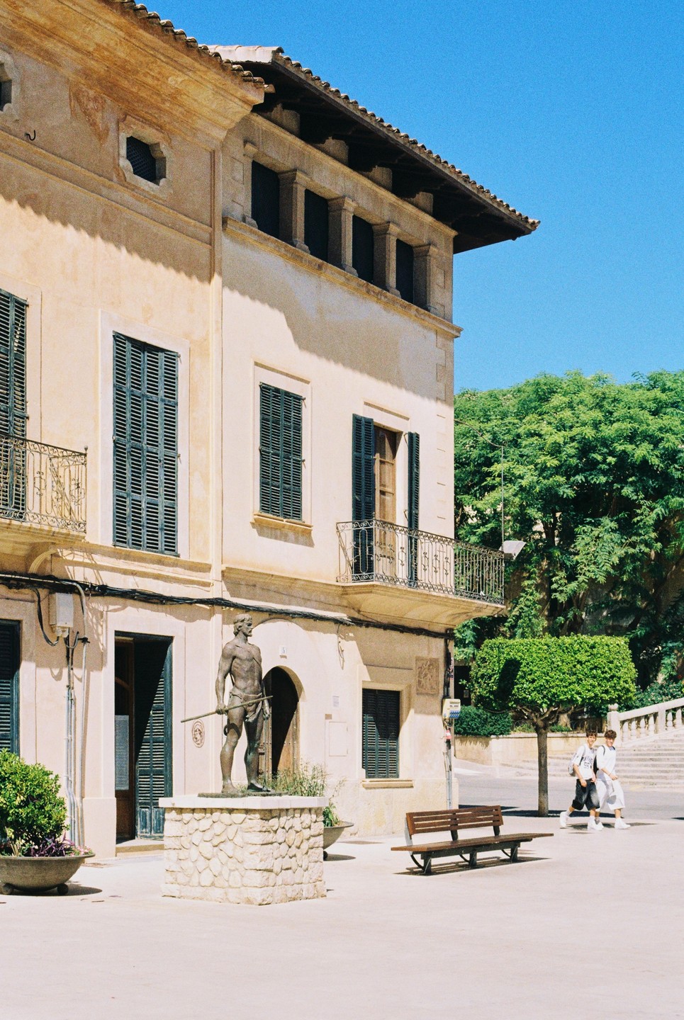 A statue of a man holding a spear stands in front of a building with green shutters and balconies, with two people walking in the background.