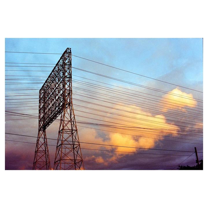 A large metal electricity pylon stands against a dramatic sky with power lines stretching across the frame.