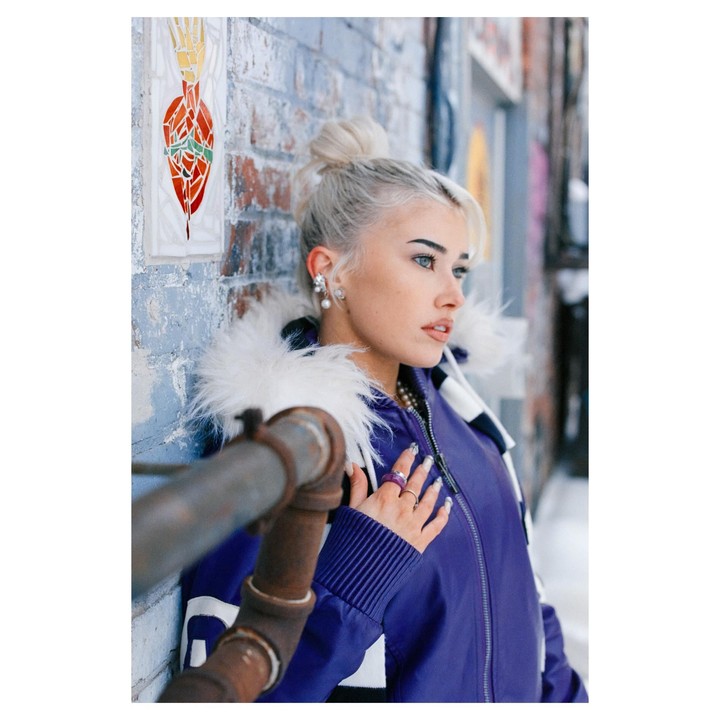 A woman with blonde hair in a purple jacket with a fur collar leans against a brick wall with a mosaic.