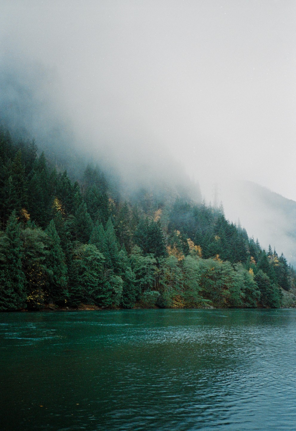 A body of water is in the foreground with a tree-covered mountain shrouded in fog in the background.