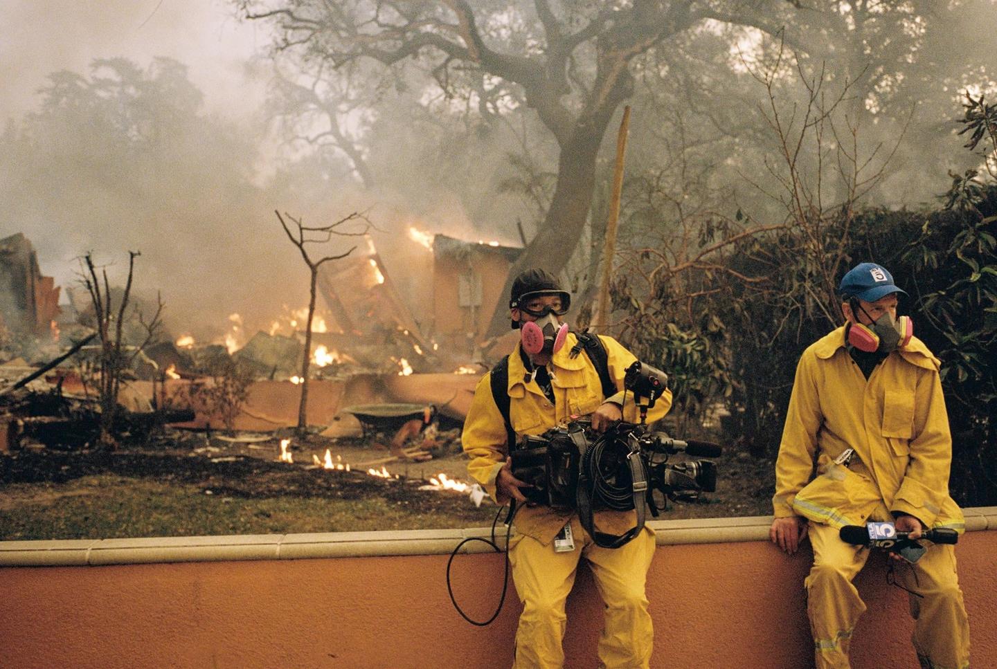Two men in yellow suits and respirators stand in front of a burning building with smoke and flames.