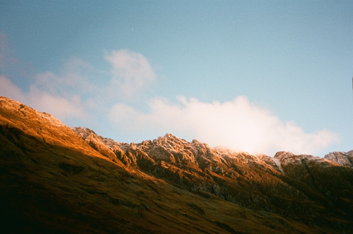 A mountain range with snow-capped peaks is illuminated by the morning sun under a clear sky with a few clouds.