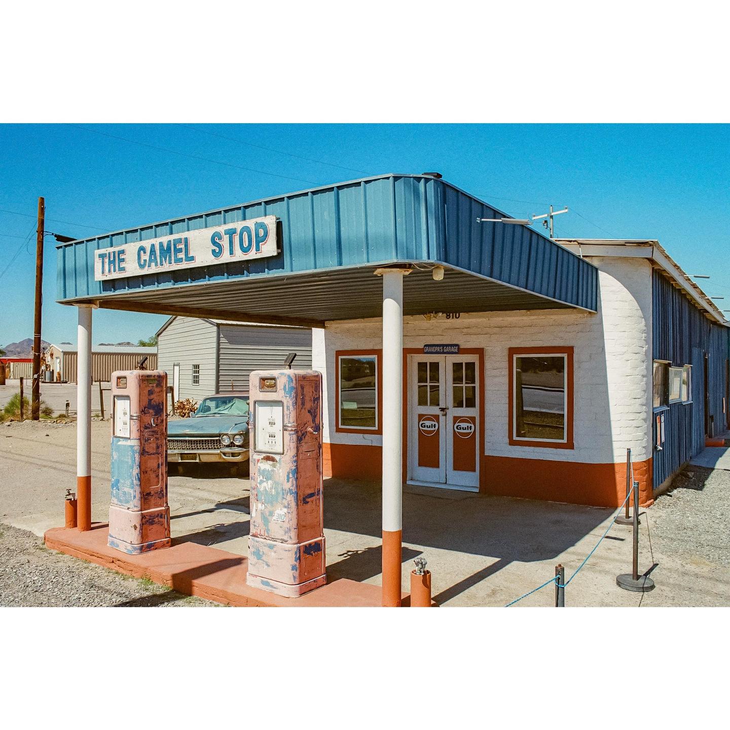An old gas station with two vintage pumps and a classic car parked behind them is visible under a clear blue sky.