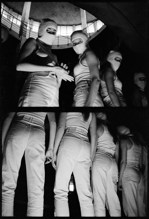 Four women in balaclavas and light-colored clothing stand in a dark, multi-level interior space.