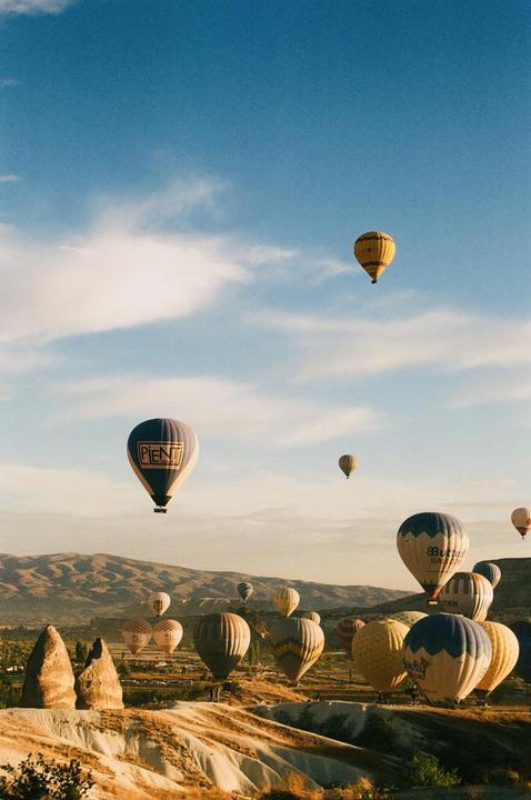Many hot air balloons float above a dry, hilly landscape with mountains in the distance under a blue sky with scattered clouds.