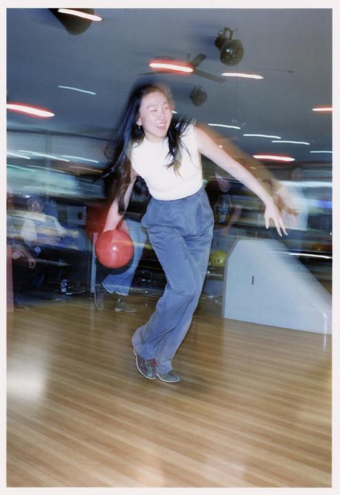 A woman smiles while bowling a red ball down a lane in a blurry bowling alley.