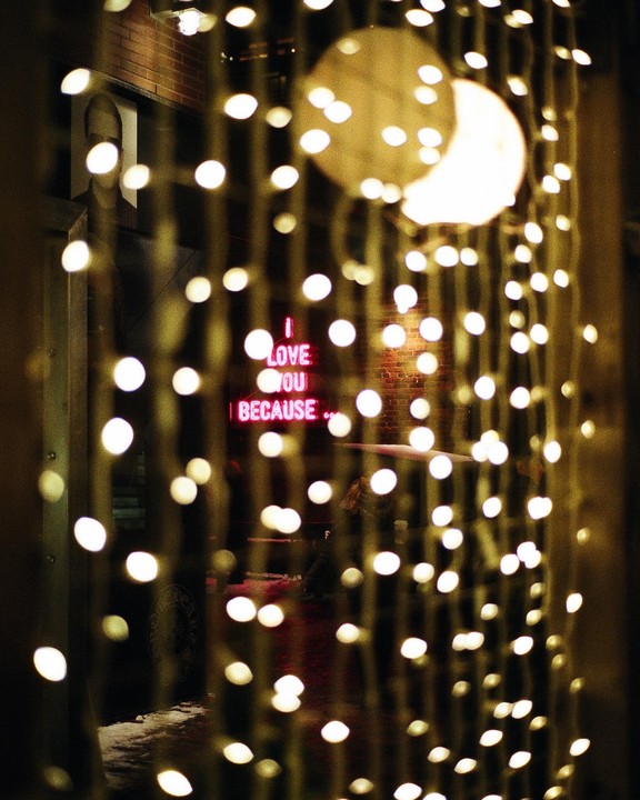 A neon sign that reads "I love you because" is visible through a curtain of string lights.