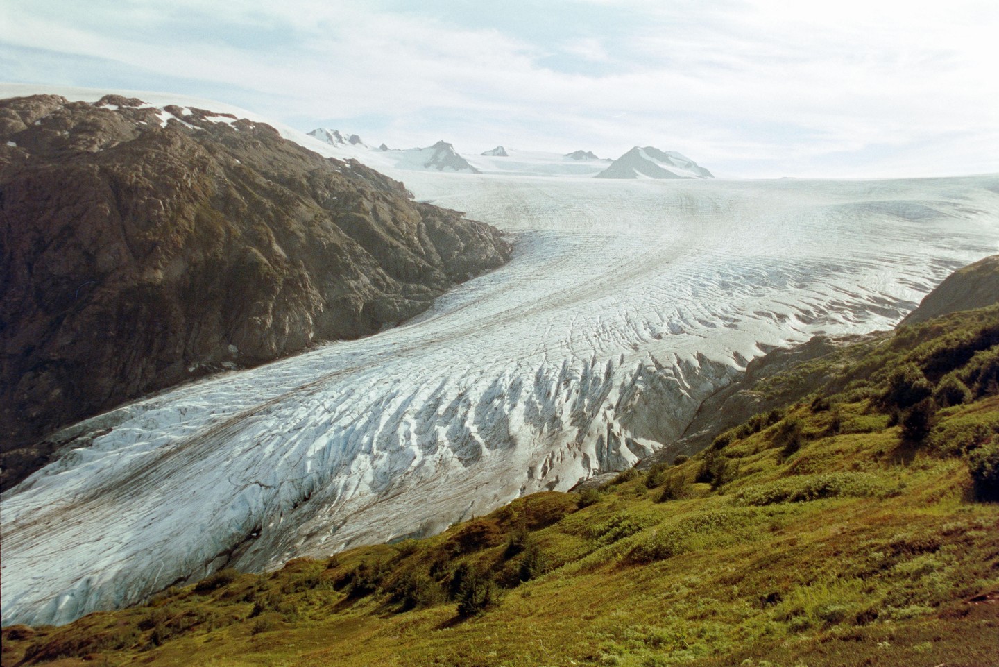A vast glacier flows between two mountainsides, with snow-capped peaks visible in the distance under a cloudy sky.