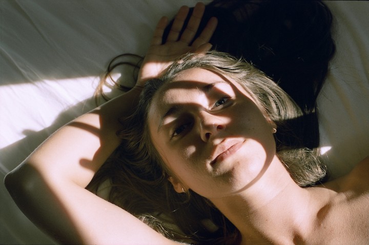 A woman lies on a bed with her arm raised, with shadows cast across her face and body.