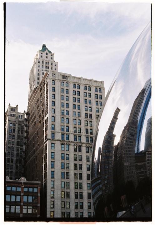 A large, reflective sculpture distorts the reflection of surrounding buildings under a bright sky.