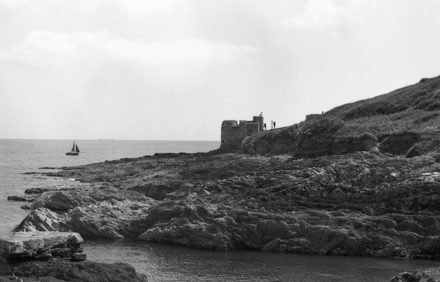 A sailboat passes by a rocky coastline with a small fort and people on a grassy hill.
