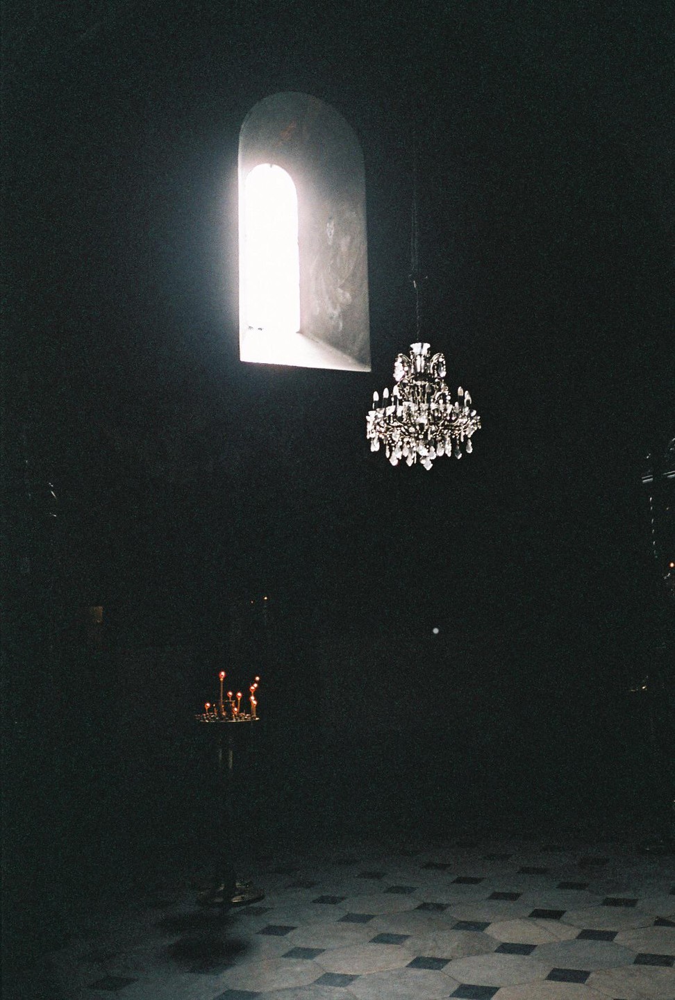 A bright arched window illuminates a dark church interior with a chandelier and lit candles.