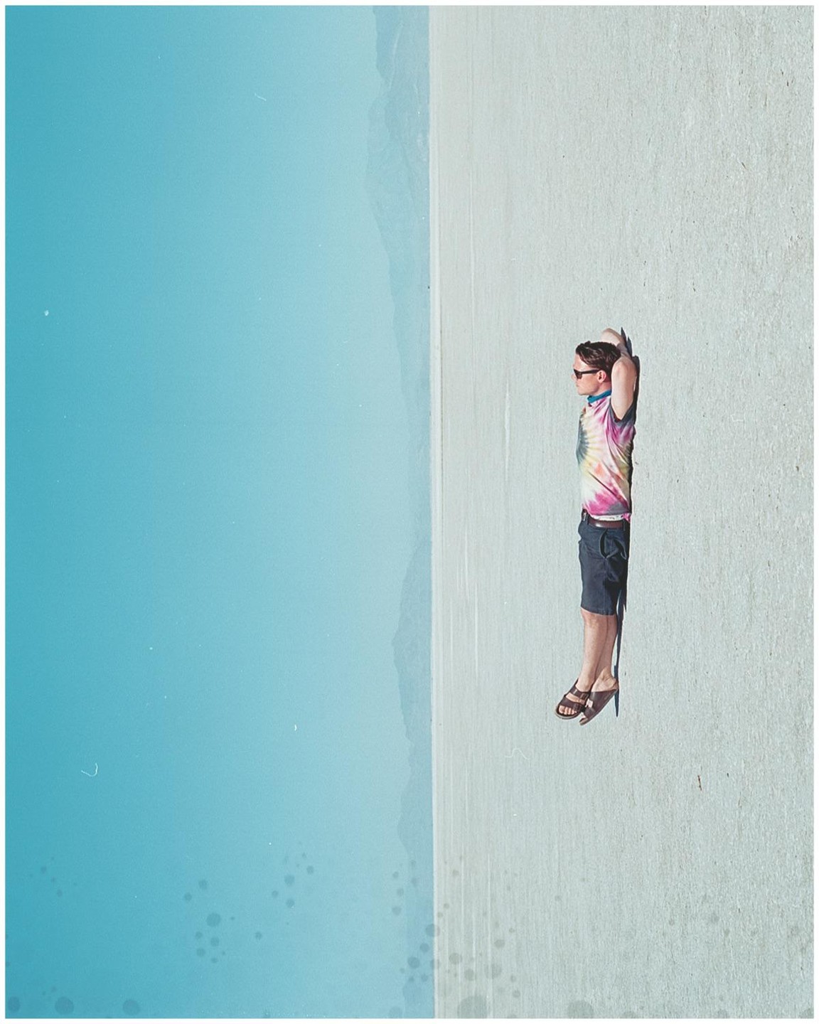 A man in a tie-dye shirt and shorts lies on a light-colored surface next to a vast expanse of blue.