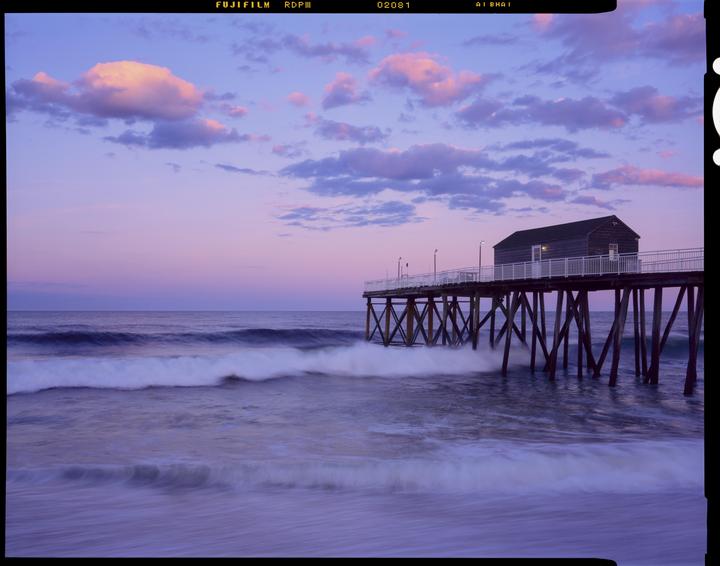 A wooden pier with a small building extends into the ocean under a purple and pink sky with clouds.