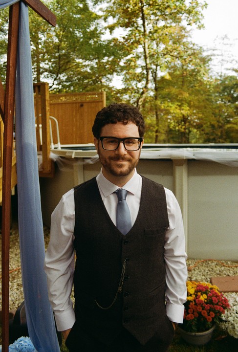 A man in a vest and tie stands smiling outdoors with trees and a pool in the background.