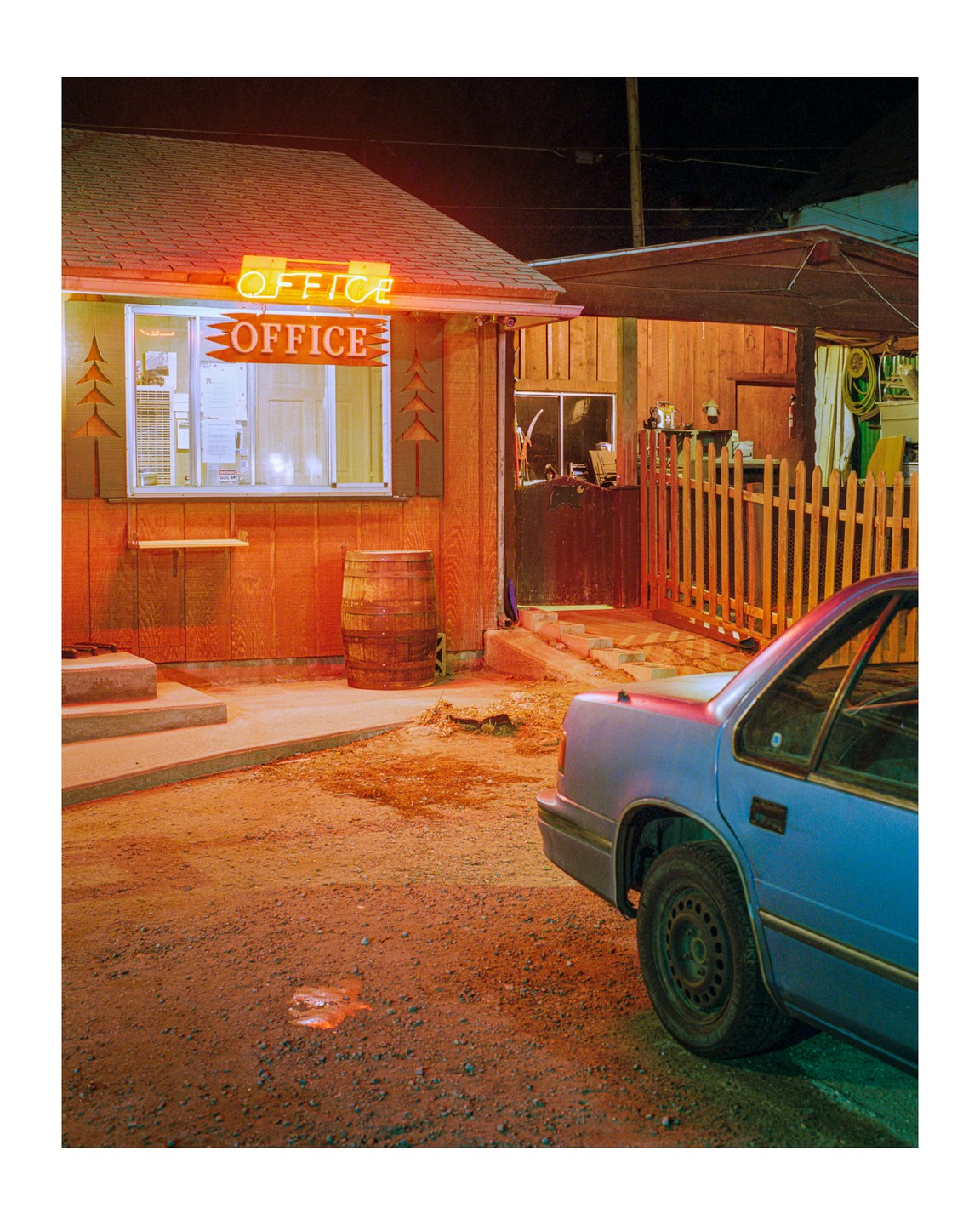 A blue car is parked on a dirt lot next to a small wooden building with a neon "OFFICE" sign at night.