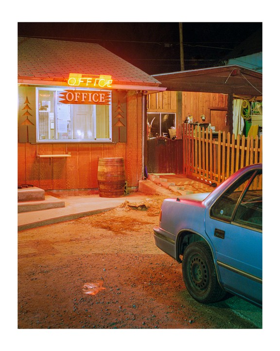 A blue car is parked on a dirt lot next to a small wooden building with a neon "OFFICE" sign at night.