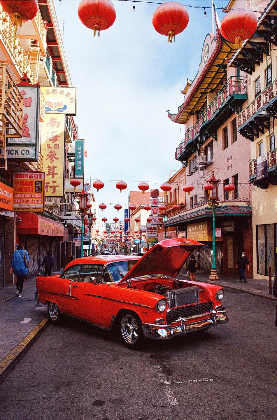A red classic car with its hood open is parked on a street lined with buildings and red lanterns.