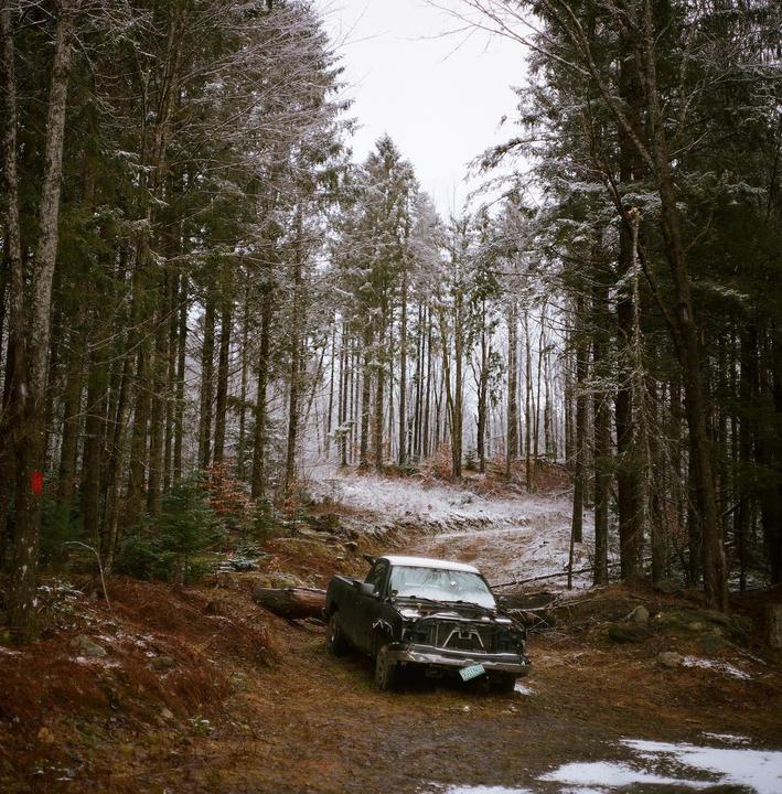 A broken-down truck sits on a muddy road in a snowy forest.