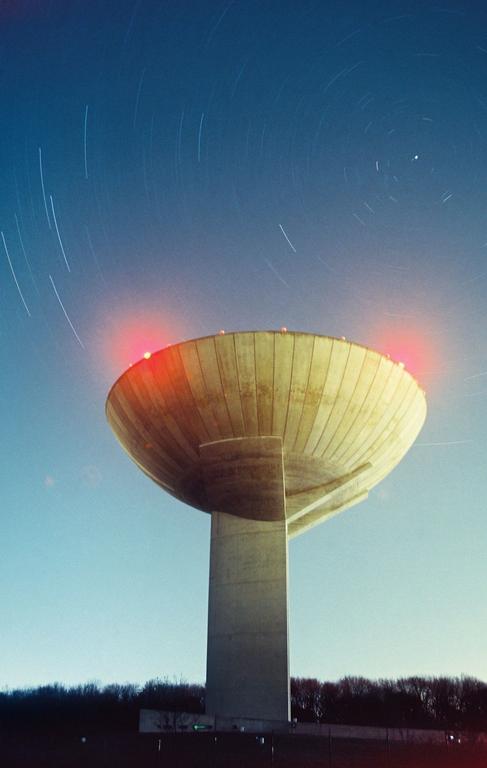 A concrete water tower with red lights on its rim stands against a blue sky with star trails.