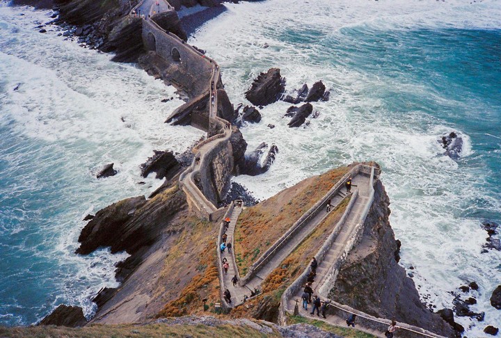 A winding stone path with people walking on it connects a rocky island to the mainland, with foamy ocean waves crashing against the shore.