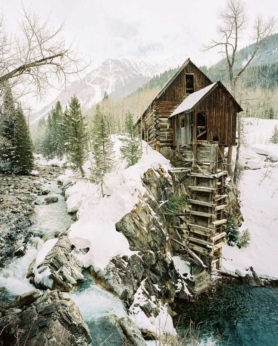 A wooden mill stands on a snowy cliff overlooking a river with mountains and trees in the background.