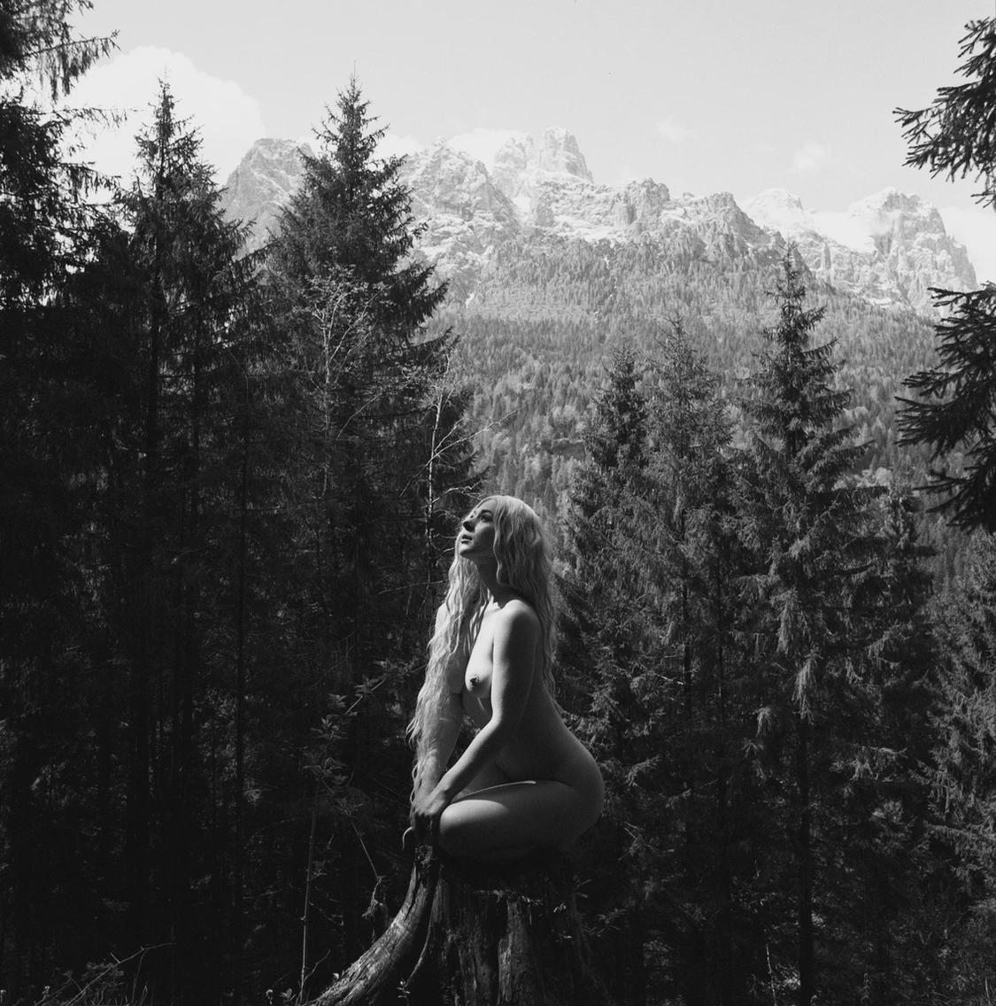 A nude woman with long hair sits on a tree stump in a forest with mountains in the background.
