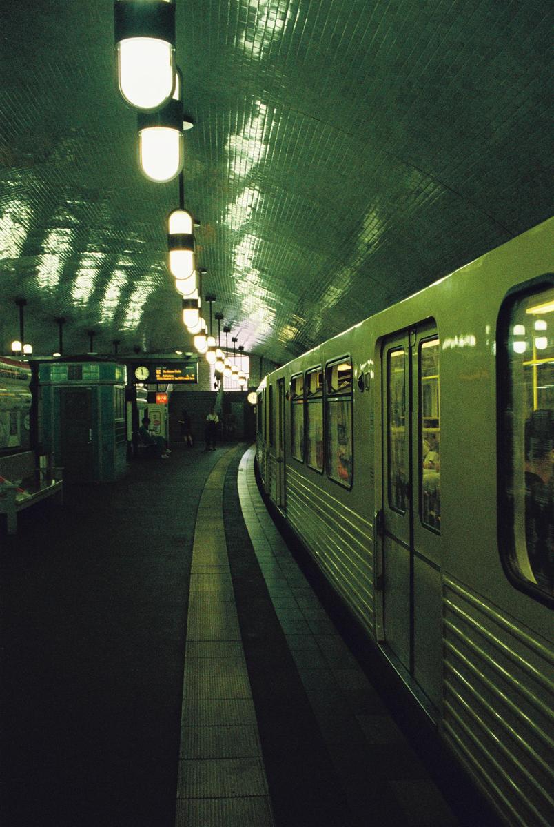 A subway train is stopped at a station platform with a curved, tiled ceiling and bright overhead lights.