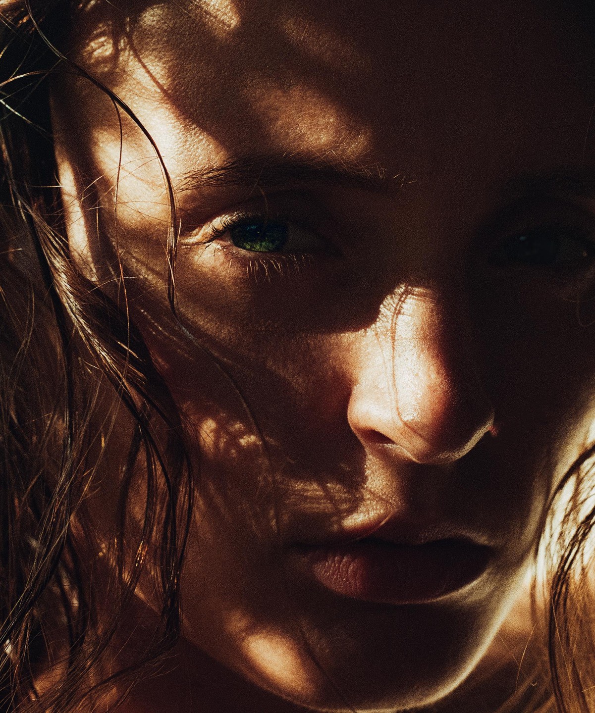 A woman's face is partially illuminated by sunlight filtering through a pattern, highlighting her green eye and wet hair.