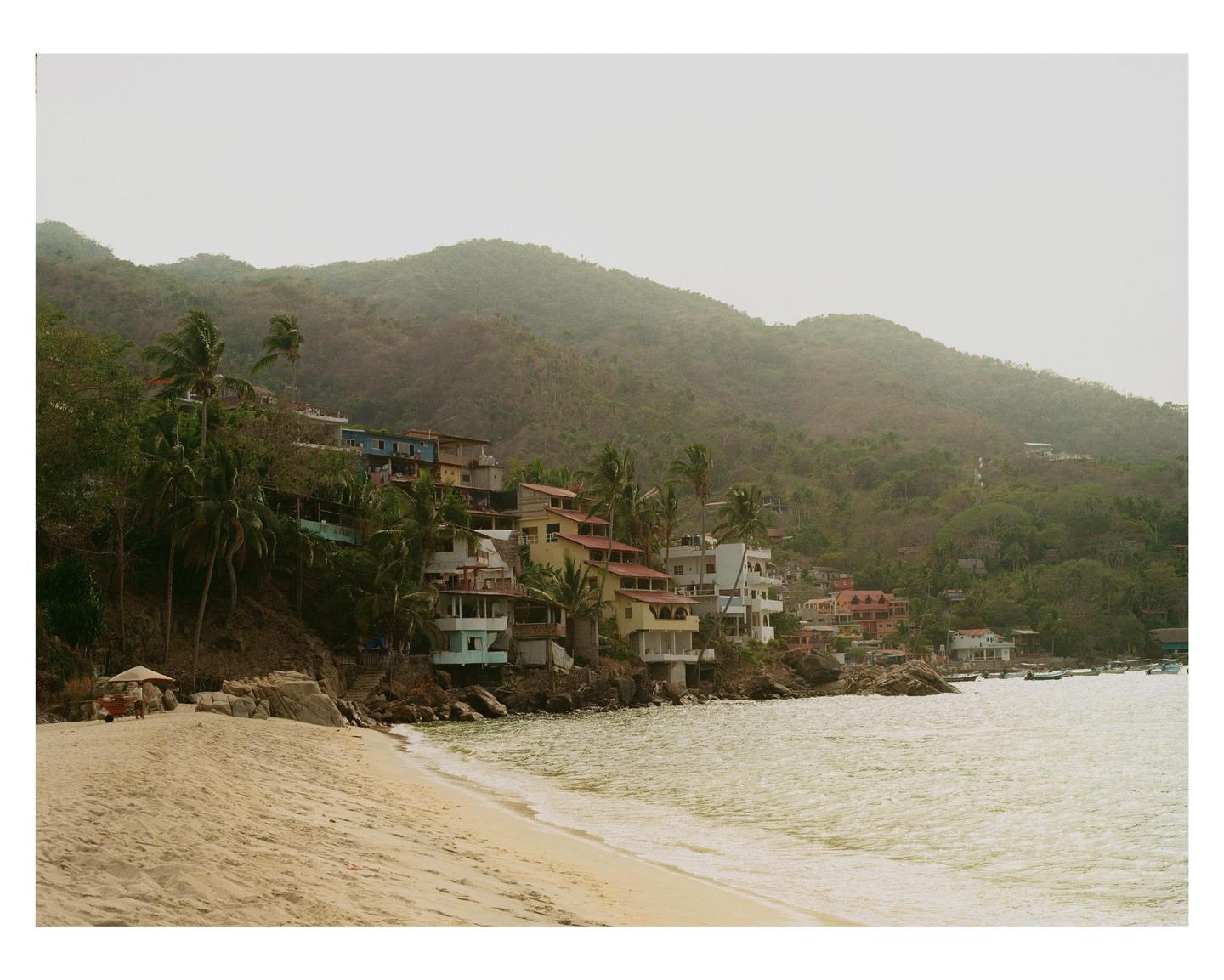 A sandy beach and ocean are in the foreground, with colorful houses built into a lush green hillside in the background.