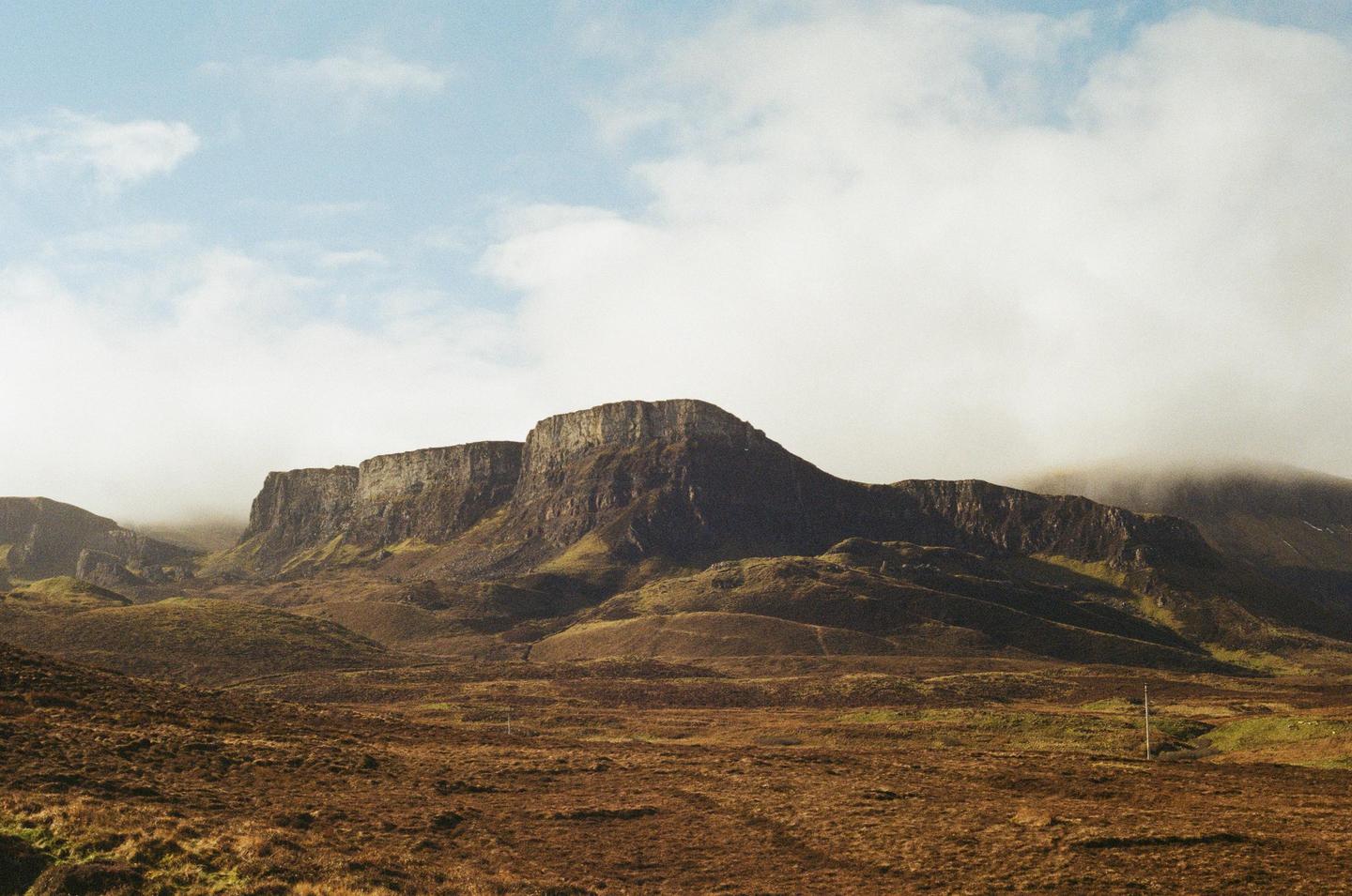 A vast, rugged landscape features a large mountain range under a cloudy sky.