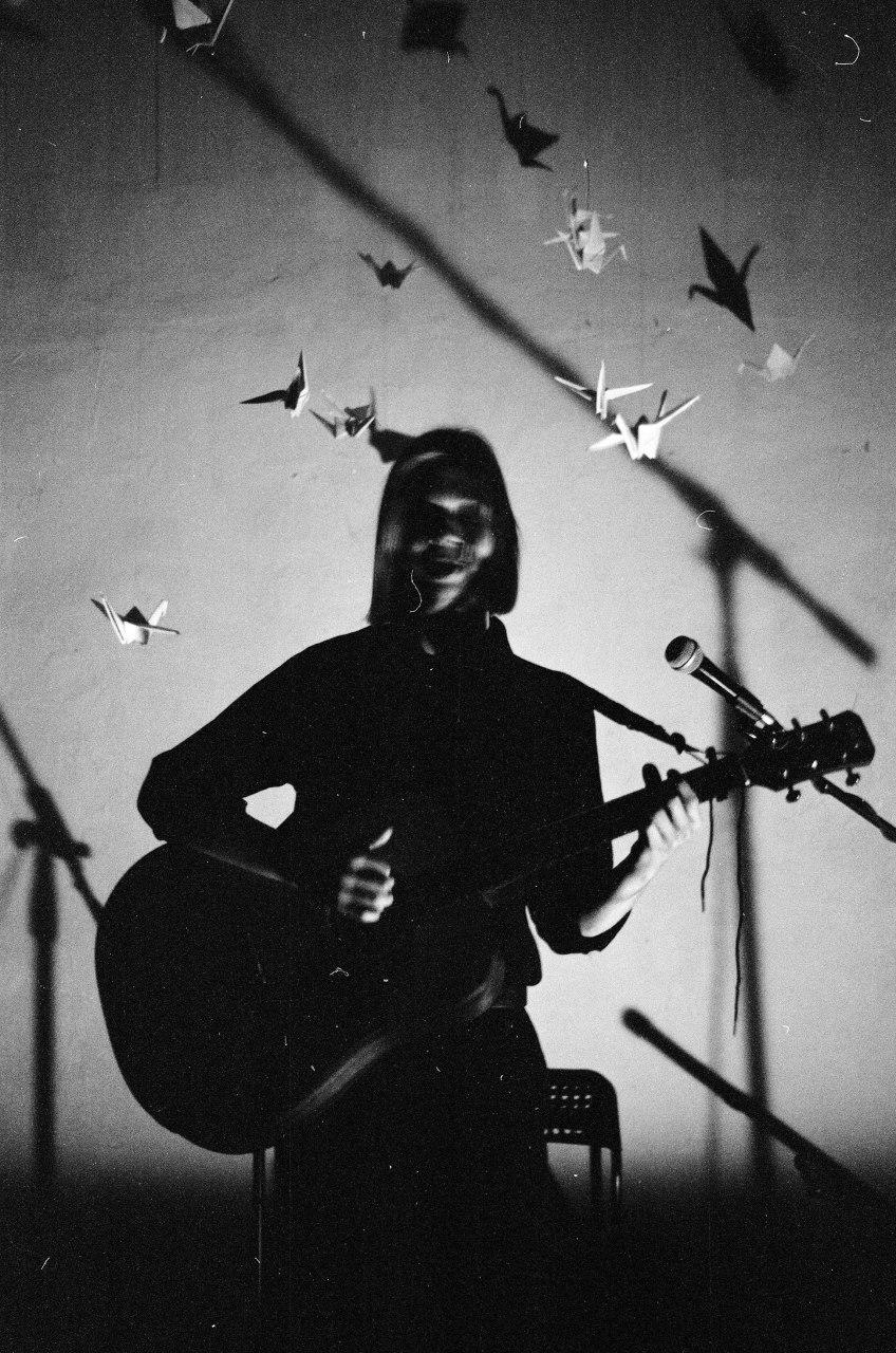 A woman plays an acoustic guitar on a stage with origami cranes hanging above her.