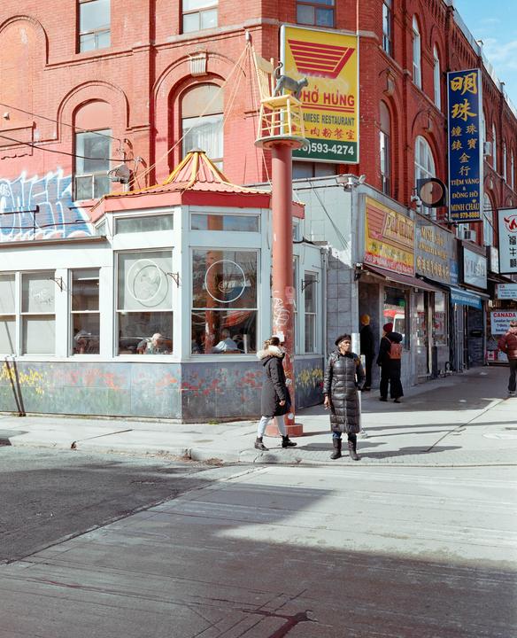 A woman in a long coat stands on a street corner in front of a brick building with storefronts and signs.