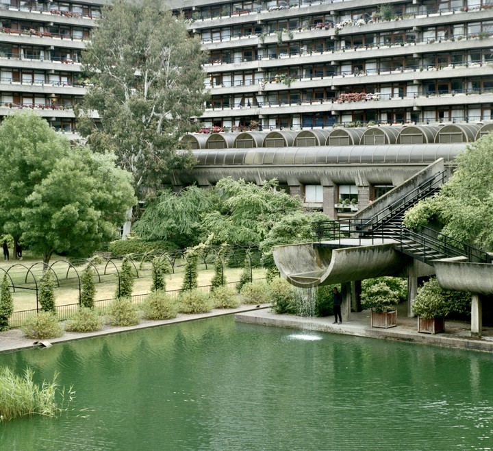 A person stands under a concrete waterfall next to a green pond, with a large building and trees in the background.