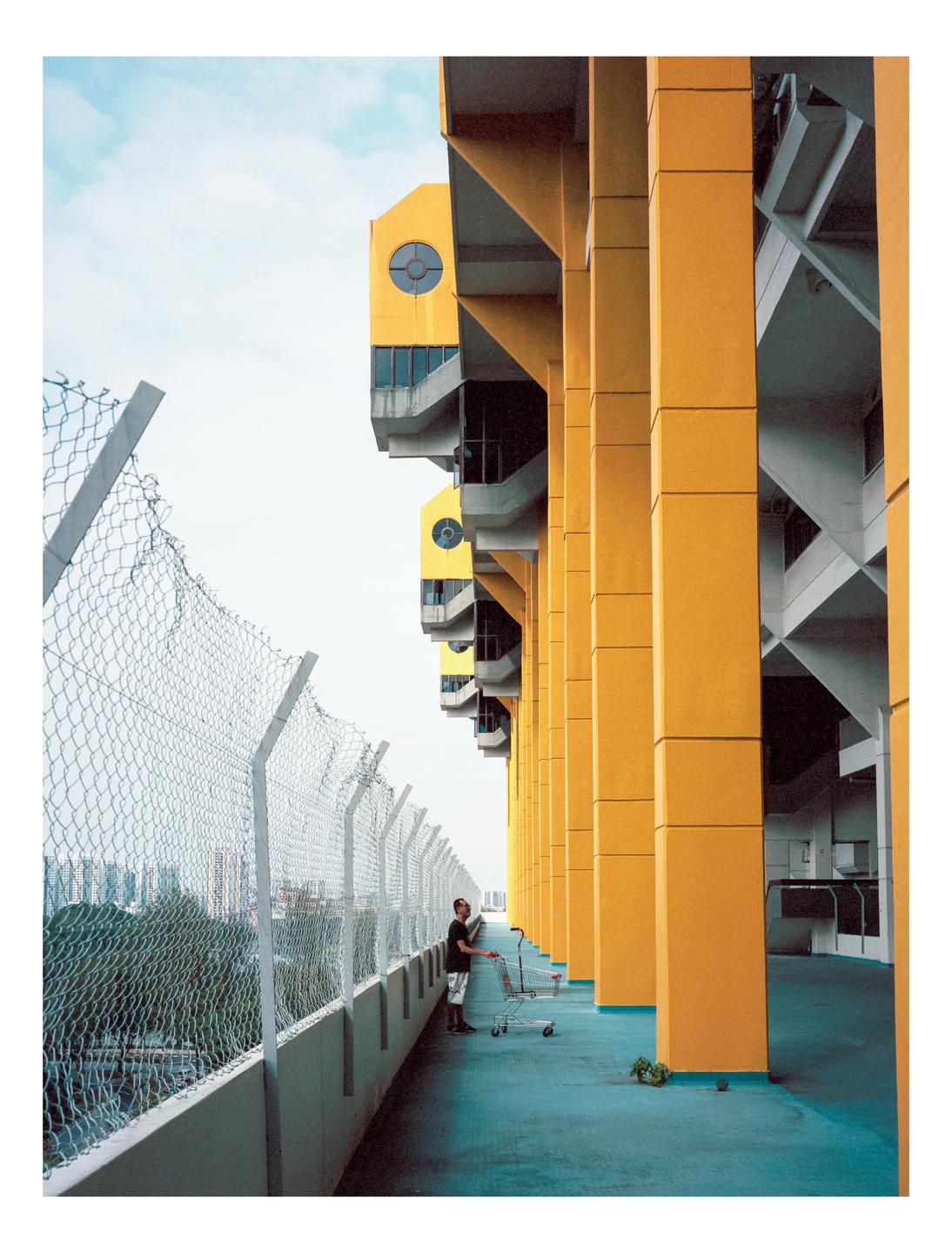 A man stands with a shopping cart on a blue walkway next to a fence and a tall yellow building.