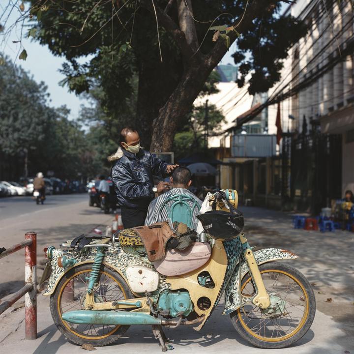 A man wearing a face mask cuts another man's hair on a street next to a colorful motorcycle.
