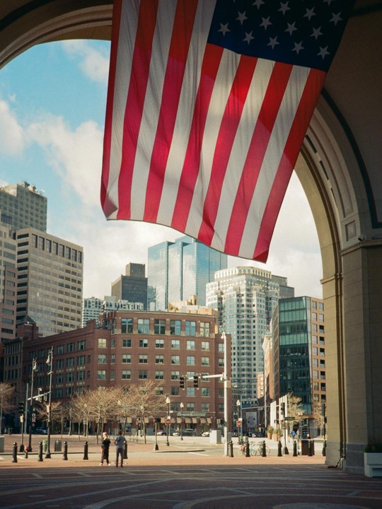 A large American flag hangs from an archway overlooking a city street with buildings and two people.