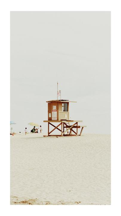 A wooden lifeguard station stands on a sandy beach with people and umbrellas in the distance.