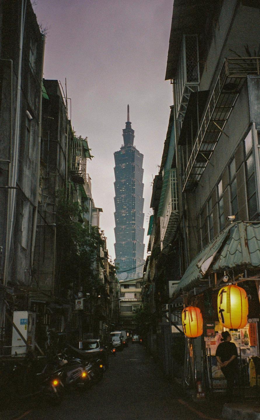 A tall skyscraper rises above a narrow street lined with buildings and parked motorcycles, with a person standing near glowing lanterns on the right.