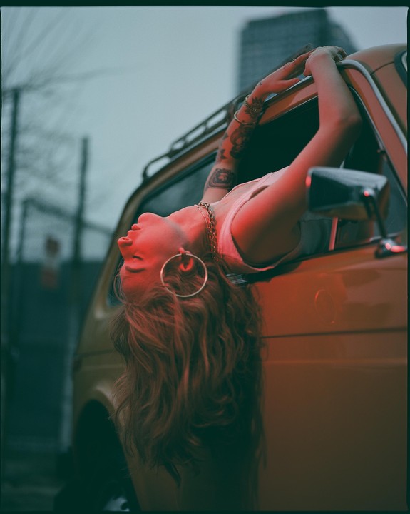 A woman with long hair and tattoos leans out of a car window, illuminated by red light.