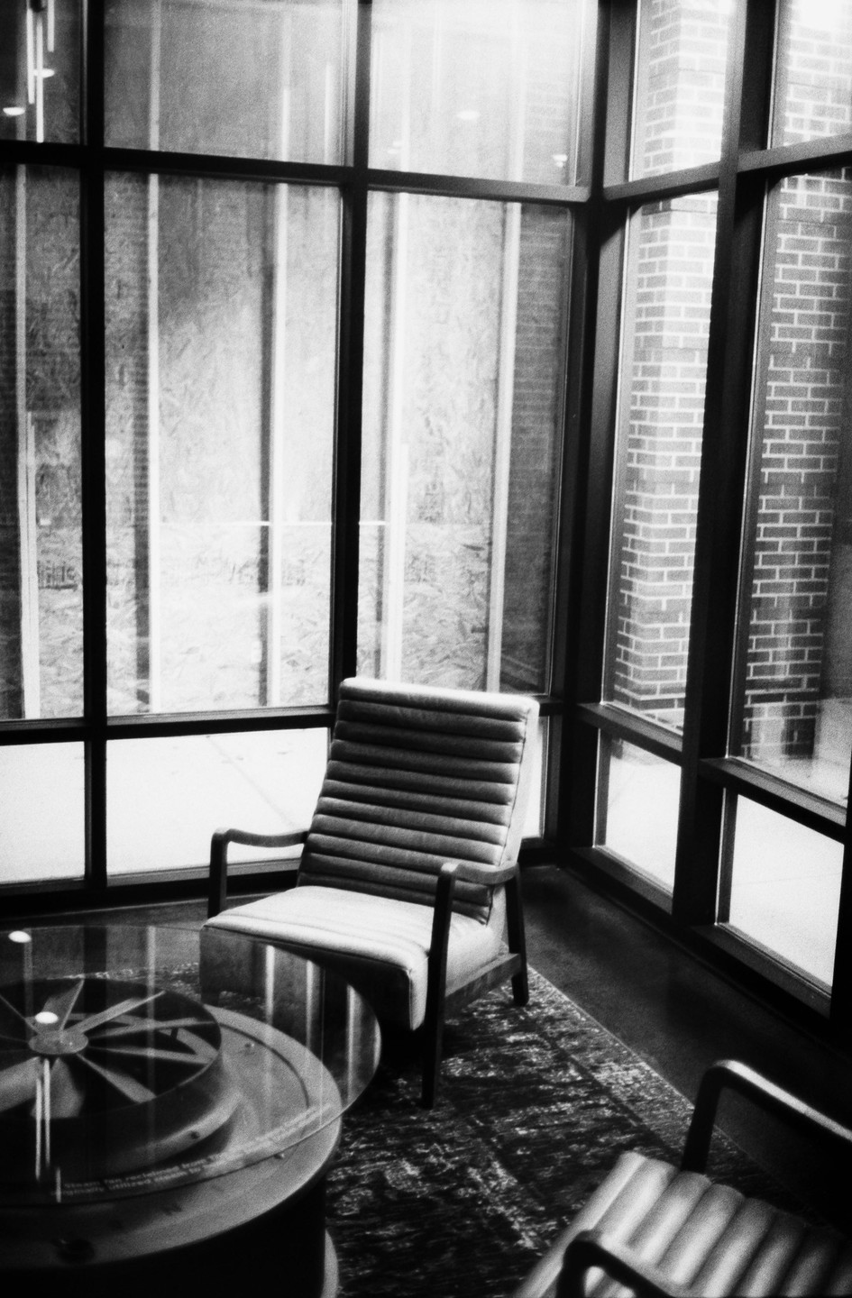 A chair sits next to a glass table in a room with large windows looking out onto a brick building.