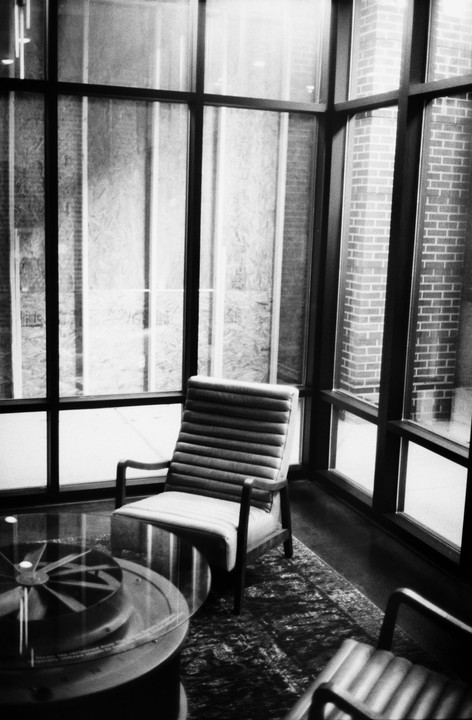 A chair sits next to a glass table in a room with large windows looking out onto a brick building.