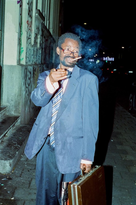 A man in a suit and tie smokes a cigarette on a street at night.