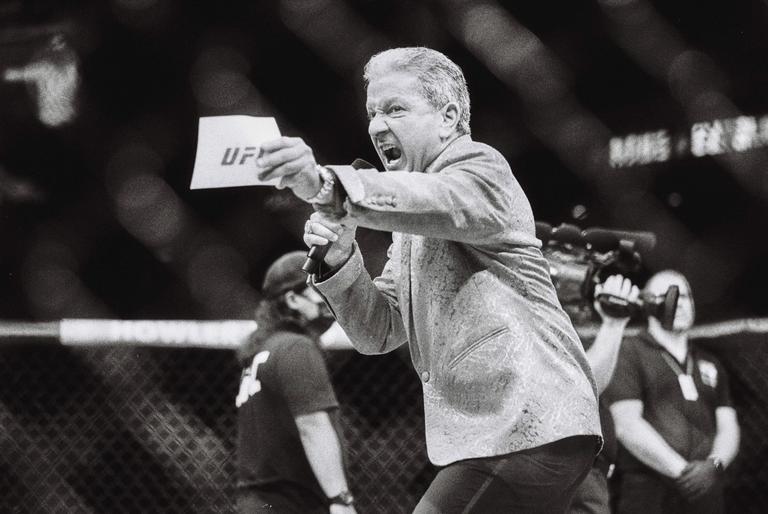 A man in a patterned jacket shouts into a microphone while holding a piece of paper with "UF" written on it.