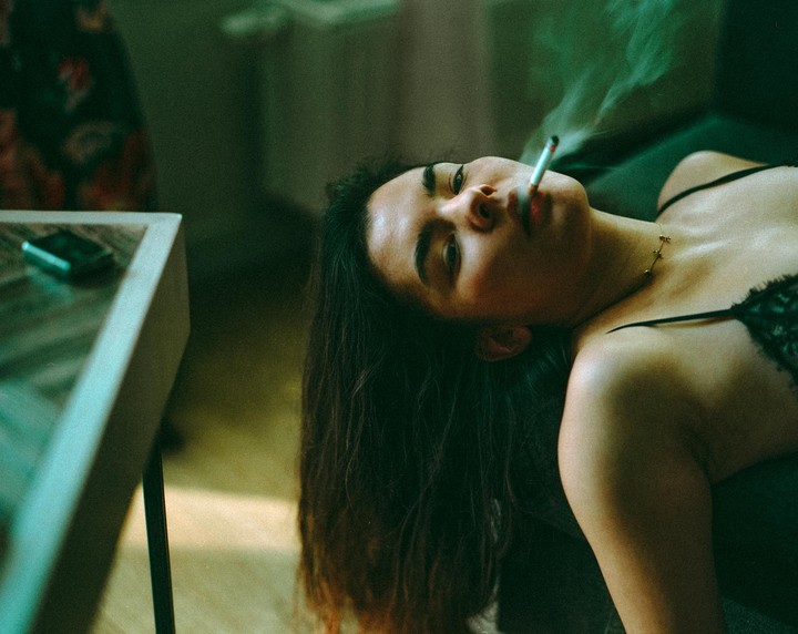 A woman with long dark hair lies on her back, smoking a cigarette with smoke rising from it.