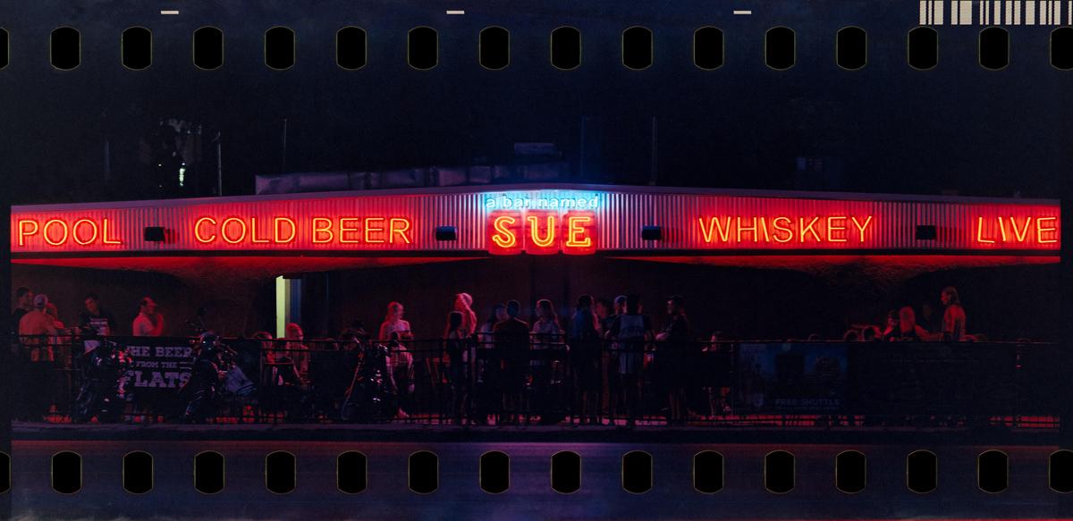 A bar with neon signs for "Pool", "Cold Beer", "Sue", "Whiskey", and "Live" is illuminated at night, with people gathered below.