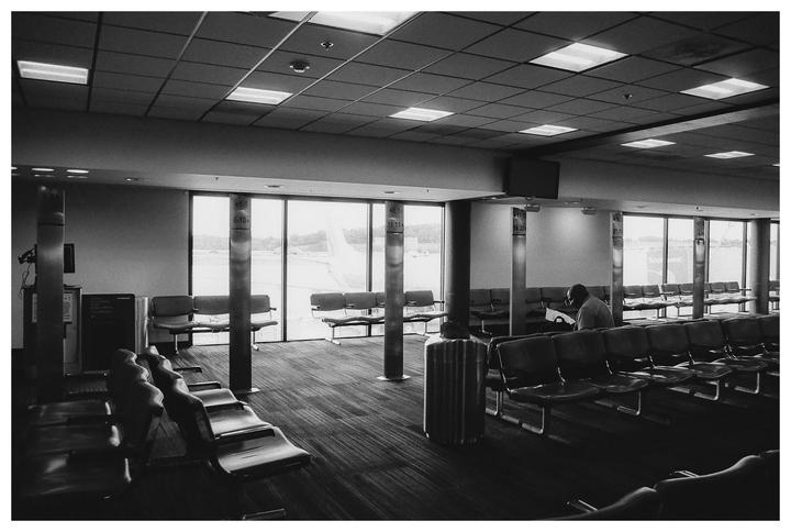 A man sits alone in an airport waiting area with rows of empty seats and large windows looking out onto an airfield.